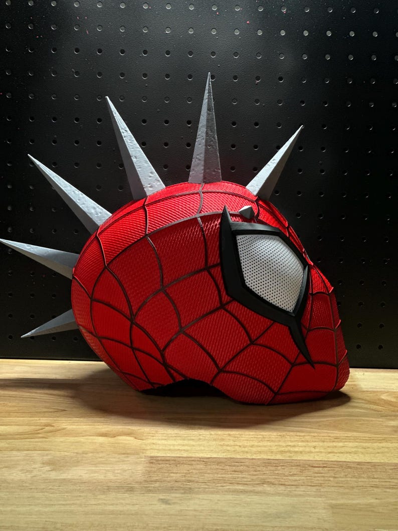 May include: A red Spider-Man mask with a web pattern and a white eye. The mask has silver spikes around the head. The mask is on a black surface.