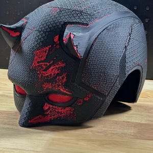 Puede incluir: Un casco negro texturizado con dos cuernos prominentes en la parte superior y acentos de color rojo vibrante en la superficie. Las aberturas para los ojos están cubiertas con una malla roja fina, con un diseño general robusto y estratificado.