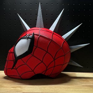 May include: A red and black Spider-Man mask with a web pattern, featuring gray spikes protruding from the top. The mask has a white mesh eye covering and is set against a black background.