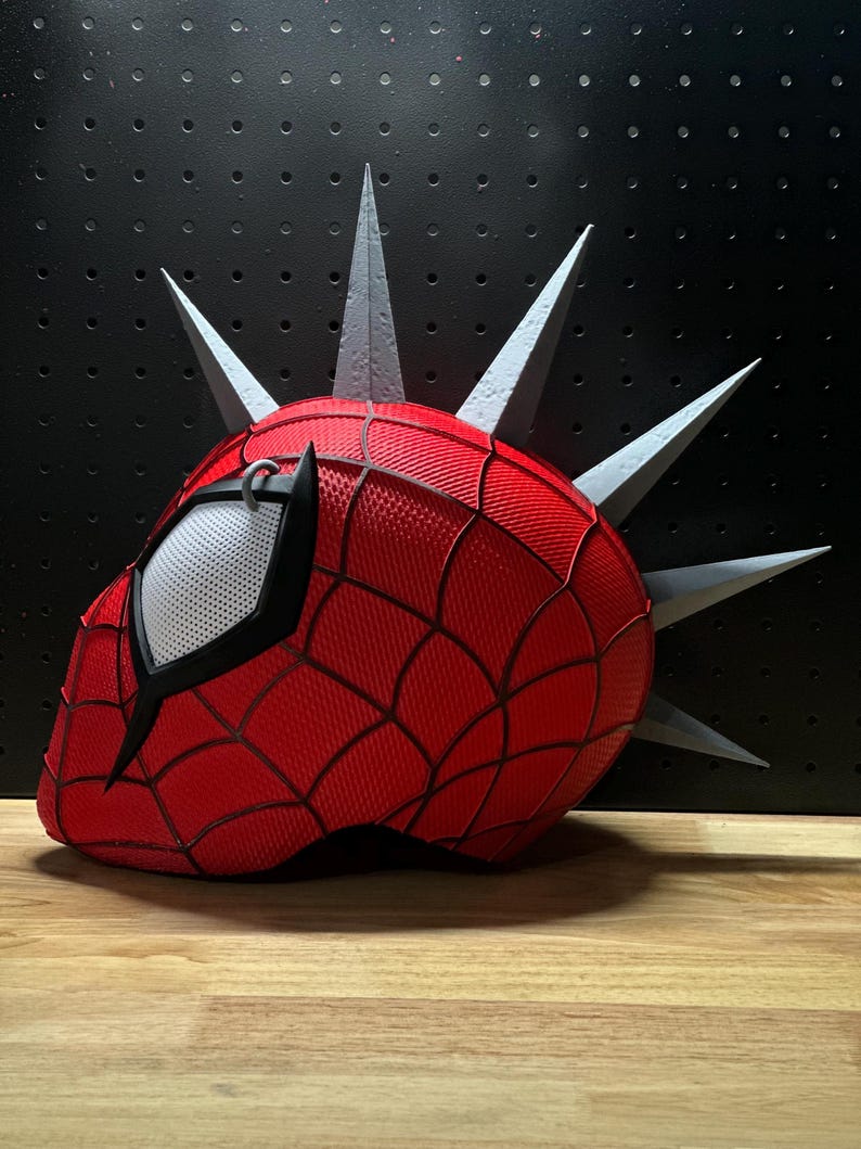 May include: A red and black Spider-Man mask with a web pattern, featuring gray spikes protruding from the top. The mask has a white mesh eye covering and is set against a black background.