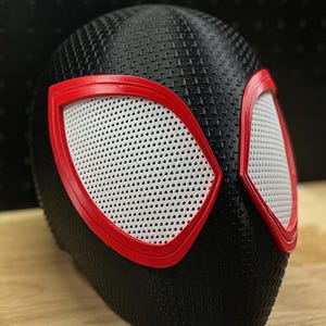May include: A black full-head mask featuring a textured surface pattern. The mask includes two large, almond-shaped eye openings outlined in bright red frames, with white mesh material covering the eye areas to allow for visibility.