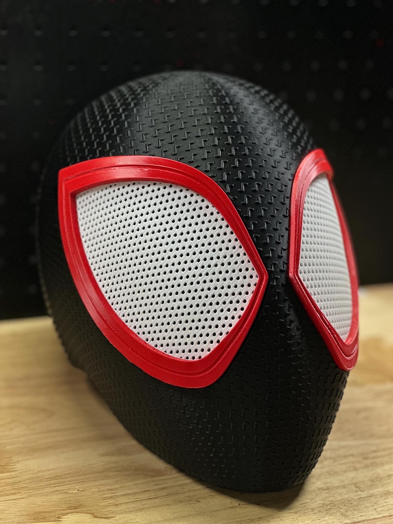 May include: A black full-head mask featuring a textured surface pattern. The mask includes two large, almond-shaped eye openings outlined in bright red frames, with white mesh material covering the eye areas to allow for visibility.