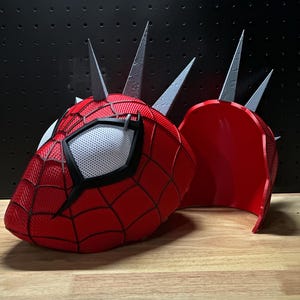 May include: A red and black Spider-Man mask with a white mesh eye area and silver spikes. The mask is split into two pieces, with the second piece resting next to the main mask on a wooden surface.