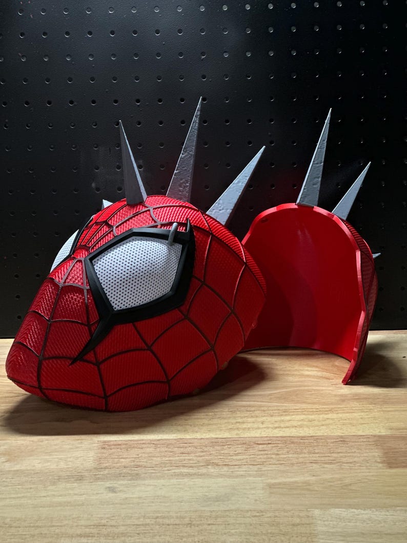 May include: A red and black Spider-Man mask with a white mesh eye area and silver spikes. The mask is split into two pieces, with the second piece resting next to the main mask on a wooden surface.