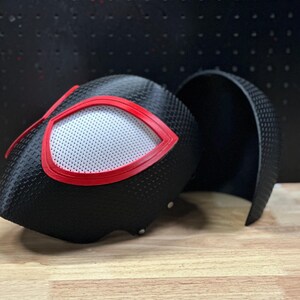 May include: A black textured helmet with a carbon fiber-like pattern, featuring large eye openings outlined in bright red. The eyes are covered with a white mesh material. A separate matching black curved piece sits beside the main helmet structure.
