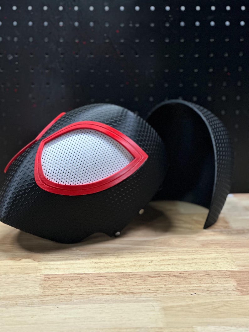 May include: A black textured helmet with a carbon fiber-like pattern, featuring large eye openings outlined in bright red. The eyes are covered with a white mesh material. A separate matching black curved piece sits beside the main helmet structure.