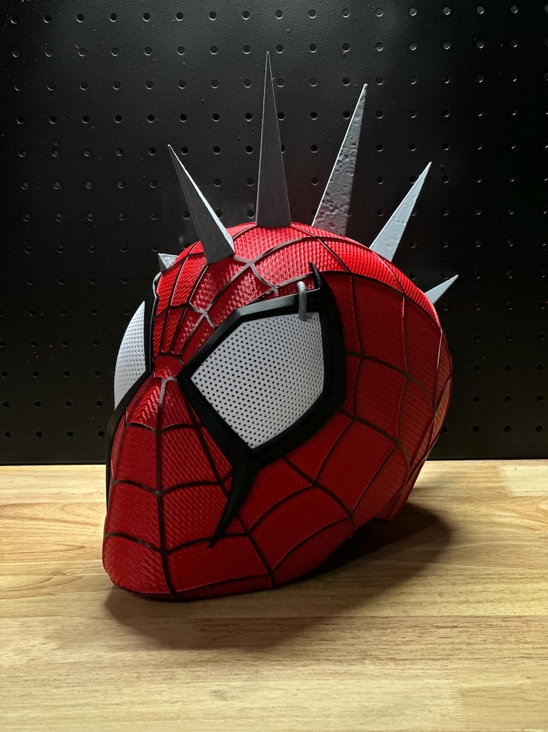 May include: A red and black Spider-Man mask with silver spikes. The mask features white mesh eye coverings and a webbed design. The mask is a cosplay item.