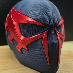 May include: A close-up of a futuristic helmet with a dark blue textured surface and striking red accents. The helmet features a distinctive design with red eye coverings and a central facial element, creating a bold and dynamic appearance. The design suggests a superhero or sci-fi theme.