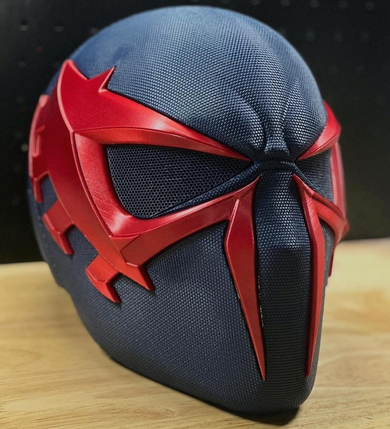 May include: A close-up of a futuristic helmet with a dark blue textured surface and striking red accents. The helmet features a distinctive design with red eye coverings and a central facial element, creating a bold and dynamic appearance. The design suggests a superhero or sci-fi theme.