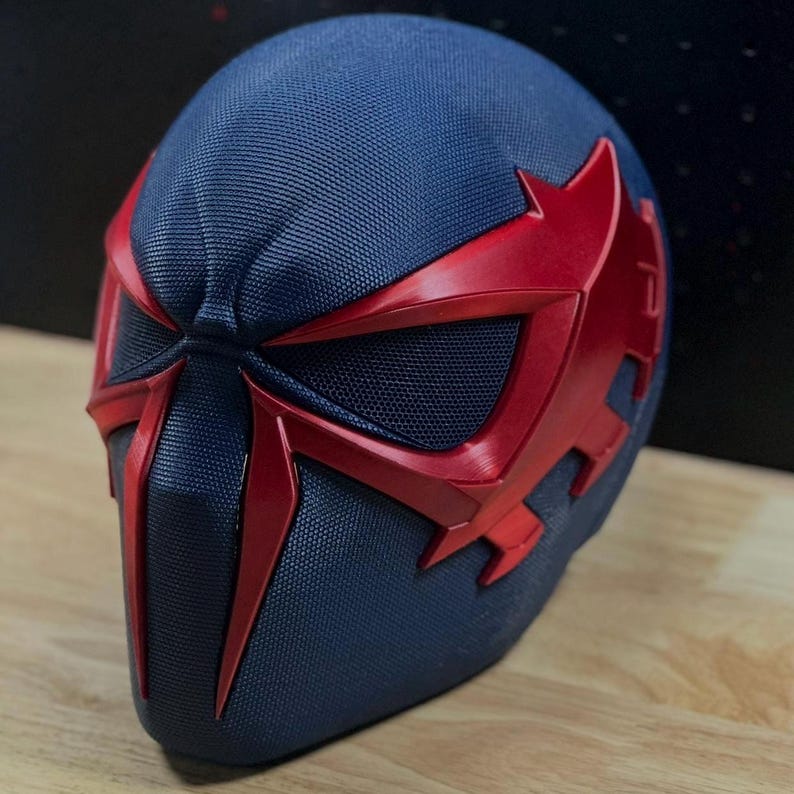 May include: A blue and red superhero mask with a textured surface. The mask features a dark blue base with red accents around the eyes and face. The design is angular and futuristic, suggesting a high-tech or comic book theme.