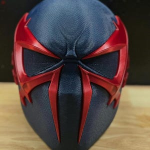 May include: A close-up of a futuristic mask in a dark blue and red color scheme. The mask has a textured surface and features angular red accents around the eyes and face, creating a striking visual contrast. The design suggests a superhero or sci-fi theme.