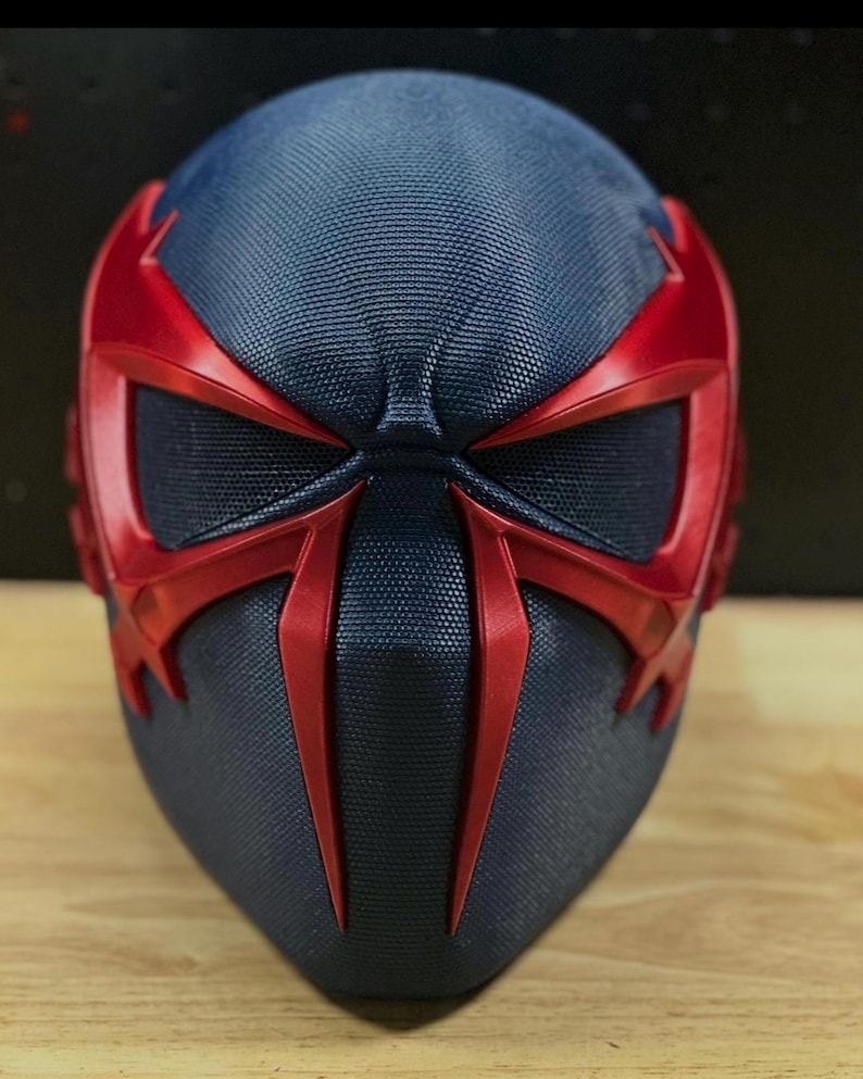 May include: A close-up of a futuristic mask in a dark blue and red color scheme. The mask has a textured surface and features angular red accents around the eyes and face, creating a striking visual contrast. The design suggests a superhero or sci-fi theme.