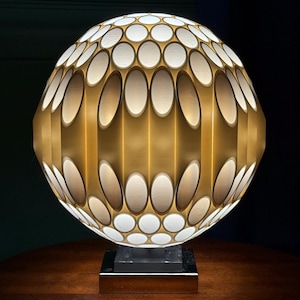 May include: A decorative table lamp with a spherical shade. The shade features a pattern of white oval and circular cutouts set against a gold-colored background. The lamp sits on a black base and a dark brown table.