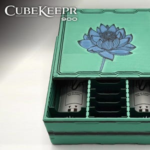 May include: A teal storage box with a blue lotus flower design on the lid. The box is labeled "CubeKeepr 900" and has internal compartments. The box has a black border and a blue logo in the bottom right corner.