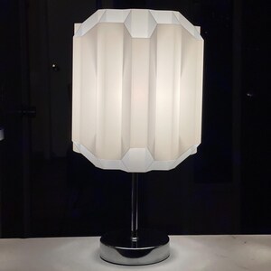 Op de afbeelding: Een witte tafellamp met een geometrische, geplooide lampenkap en een chromen voet.