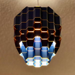 May include: A modern, geometric pendant light fixture. The lamp features a dark blue, hexagonal design with gold accents. The light emits a warm glow, creating a striking visual effect. The fixture is suspended from a single cord.