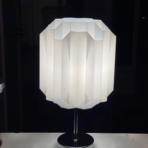 Op de afbeelding: Een witte tafellamp met een geometrische, geplooide lampenkap en een chromen voet.
