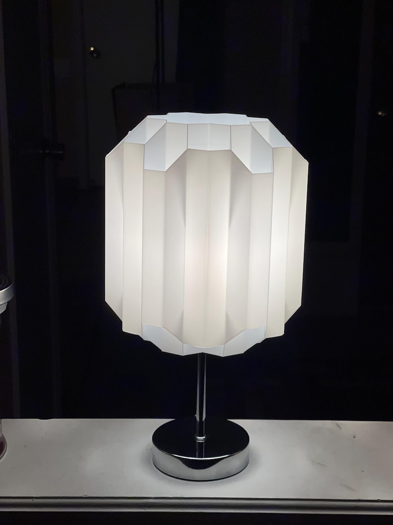 Op de afbeelding: Een witte tafellamp met een geometrische, geplooide lampenkap en een chromen voet.