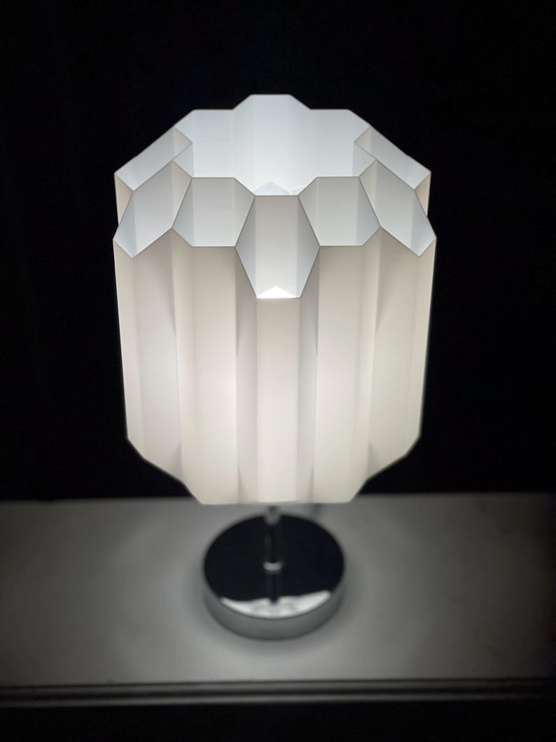 Op de afbeelding: Een witte tafellamp met een geometrische, geplooide kap en een chromen voet.