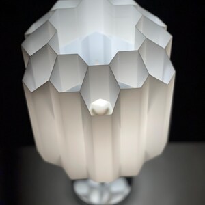 Op de afbeelding: Een witte tafellamp met een geometrische, honingraatvormige kap. De lamp is aan en het licht schijnt door de gaten in de kap.