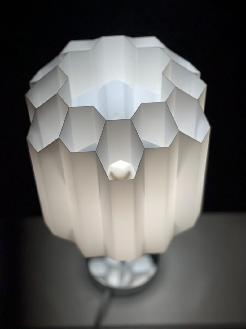 Op de afbeelding: Een witte tafellamp met een geometrische, honingraatvormige kap. De lamp is aan en het licht schijnt door de gaten in de kap.