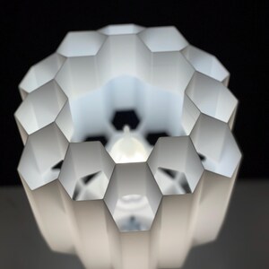 Op de afbeelding: Witte lampenkap in honingraatvorm met een witte gloeilamp erin. De lampenkap is gemaakt van dun, gevouwen plastic en heeft een geometrisch ontwerp.