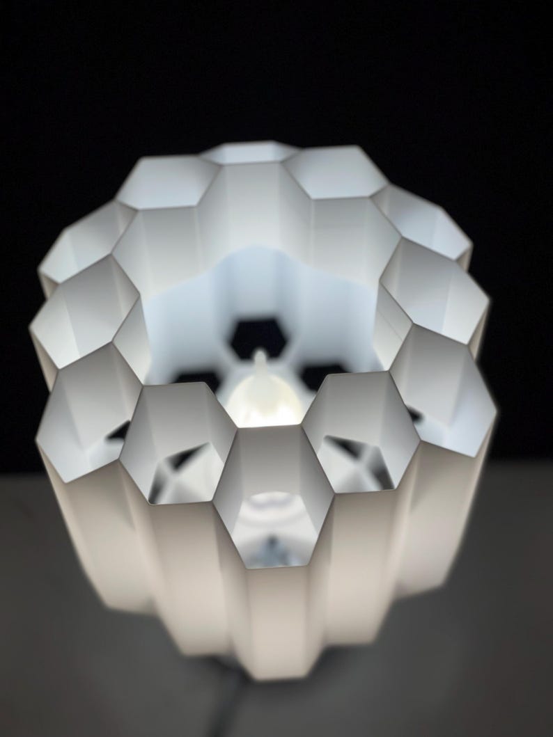 Op de afbeelding: Witte lampenkap in honingraatvorm met een witte gloeilamp erin. De lampenkap is gemaakt van dun, gevouwen plastic en heeft een geometrisch ontwerp.