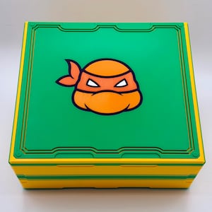 May include: A square, two-toned box with a green top and yellow sides. The top features a cartoon image of a character's face with an orange mask and a green border. The box is likely for storage or display.