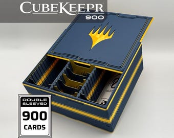 May include: A navy blue card storage box labeled CubeKeepr 900 with a yellow flame logo on the lid. The interior features yellow dividers and slots for cards. A white rectangular graphic on the side displays the text Double Sleeved 900 Cards.