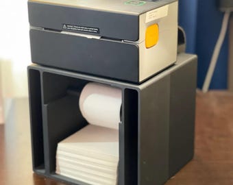 May include: A black and gray label printer with a yellow button sits atop a dark gray label holder. The label holder contains a roll of white labels and a stack of individual labels. The printer has a warning label.