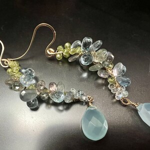May include: Gold-coloured earrings with clusters of small, multi-coloured gemstones. The earrings have a cascading design with blue, green, and clear stones, and a larger, teardrop-shaped blue stone at the bottom. The earrings are on a dark background.