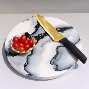 Puede incluir: Una tabla redonda para servir con un diseño de mármol blanco y gris. Un cuchillo dorado con mango negro descansa sobre la tabla. Un pequeño cuenco con borde dorado contiene tomates cherry rojos y naranjas.