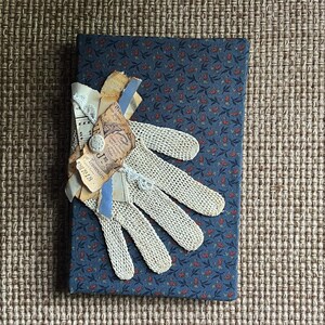 May include: A blue fabric-covered journal with a floral pattern, adorned with a vintage crochet glove and paper ephemera. The journal is embellished with torn paper, a button, and blue ribbon.