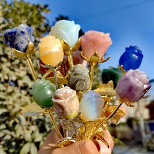 Peut inclure: Un bouquet de roses en pierres précieuses de différentes couleurs, dont le bleu, le rose, le vert et le violet. Les têtes de roses sont en pierres polies, avec des tiges et des feuilles en métal doré. L'arrangement est tenu dans une main.