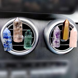 May include: An assortment of crystal point car air fresheners in various colors, including blue, brown, green, purple, yellow, and pink. The crystals are displayed in the car's air vents, showcasing their unique shapes and textures.