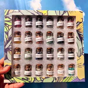 May include: A collection of twenty-four small glass jars with cork stoppers, each filled with different tumbled gemstones and labelled with names like Cherry Quartz, Citrine, Lapis Lazuli, and Turquoise, arranged in a floral-patterned box.