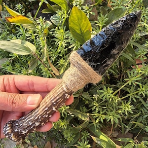 Puede incluir: Un cuchillo artesanal de obsidiana con una hoja oscura y brillante. El mango es de asta y está envuelto con hilo natural. El cuchillo se muestra sobre un fondo de follaje verde.