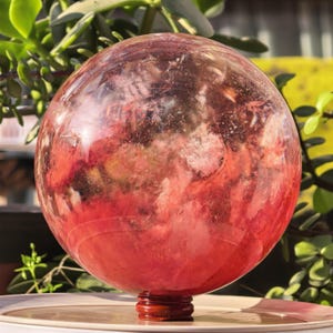 May include: A large, transparent sphere with a reddish-pink interior, set on a small, dark wooden base. The sphere's surface has a cloudy, textured appearance, with variations in colour and opacity. Green foliage forms the background.
