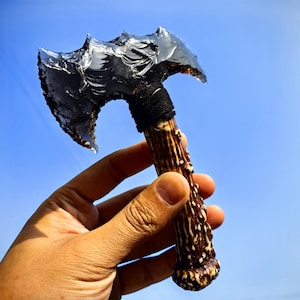 May include: A handcrafted obsidian axe with a dark, reflective blade. The handle is made of brown antler, wrapped with black material. The axe is held against a clear blue sky.
