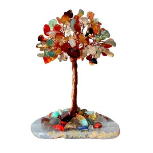 May include: A decorative tree sculpture with a copper-colored wire trunk and branches. The branches are adorned with colorful, polished gemstones in shades of red, orange, green, and white. The tree sits on a gray, stone-like base, also decorated with gemstones.