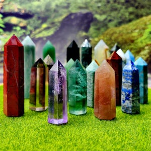 May include: A selection of gemstone towers in colours including red, green, purple, and blue. The crystals are displayed on a bed of green grass, with a blurred natural backdrop. Each tower has a pointed top.
