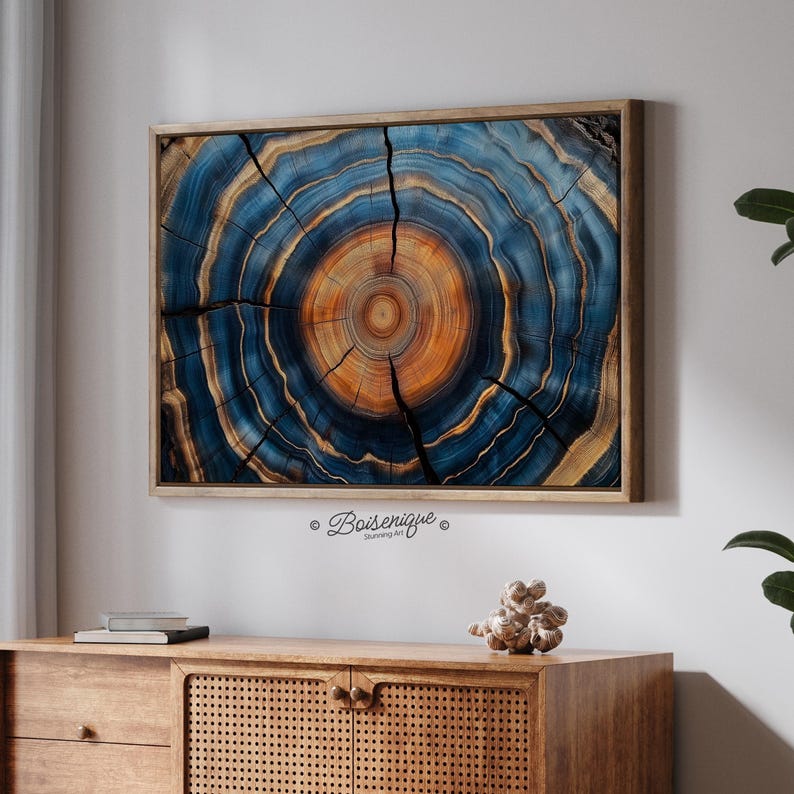 May include: A framed wood slice art piece with a detailed cross-section design. The artwork features concentric rings in shades of blue, brown, and orange, with natural cracks. The piece is mounted in a wooden frame and hangs above a wooden cabinet.