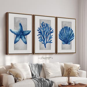 Op de afbeelding: Set van drie ingelijste wandkunstprints met aquarel illustraties van een blauwe zeester, koraal en zeeschelp. Elke afbeelding staat tegen een grijze achtergrond in een gouden lijst, wat een kustthema creëert.
