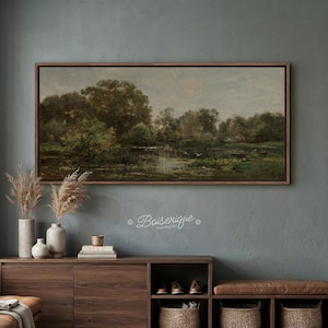 May include: A rectangular landscape painting in a dark wood frame depicting a calm pond surrounded by dense green trees and foliage under a muted sky. The artwork features a small boat on the water and is displayed above a wooden cabinet.