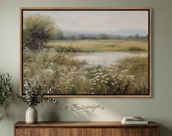 May include: A framed landscape painting depicts a serene meadow scene with a body of water, wildflowers, and distant hills. The artwork is in a wooden frame and hangs above a wooden cabinet. The painting has a soft color palette of greens, yellows, and blues.