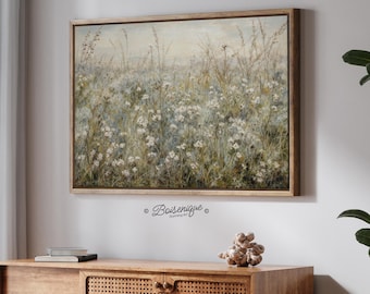 May include: A framed landscape painting featuring a field of white wildflowers and tall grasses. The artwork has a muted color palette with shades of green, blue, and beige. The frame is made of light-colored wood. The painting is hanging above a wooden cabinet.