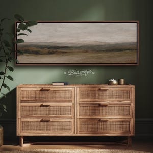 May include: A landscape painting in a dark brown frame hangs above a wooden dresser with six drawers. The painting depicts a muted scene of rolling hills and a cloudy sky. The dresser has a woven rattan design on the drawer fronts.