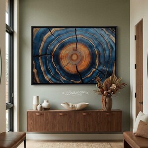 May include: A framed artwork featuring a close-up of a tree cross-section. The artwork displays concentric rings in shades of blue, brown, and orange, with visible cracks. The piece is mounted on a light green wall above a wooden cabinet.