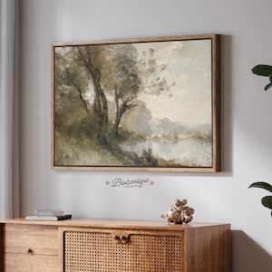 Puede incluir: Pintura de paisaje enmarcada que representa una serena escena junto al agua. La obra de arte presenta tonos apagados de verde, marrón y beige, con árboles bordeando el agua. La pintura se muestra en un marco de madera natural, lo que aumenta su encanto rústico.