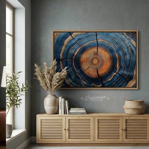 May include: A framed artwork featuring a cross-section of a tree trunk, showcasing concentric rings in shades of blue, orange, and brown. The artwork is displayed above a wooden cabinet with a vase of dried plants, books, and a woven basket.
